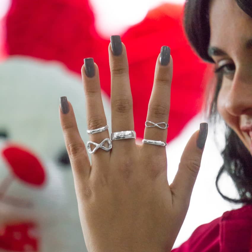 Mano femenina mostrando una selección de anillos de plata con diseños de infinito y alianzas de plata, con un fondo desenfocado de corazón rojo, simbolizando amor y compromiso.