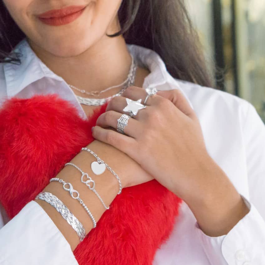 Mujer abrazando un cojín de corazón y luciendo algunas joyas de plata, incluyendo pulseras trenzadas, con corazones e infinitos y anillos con diseños de estrella, clave de sol y solitario de circonita, perfectos para regalo de San Valentín