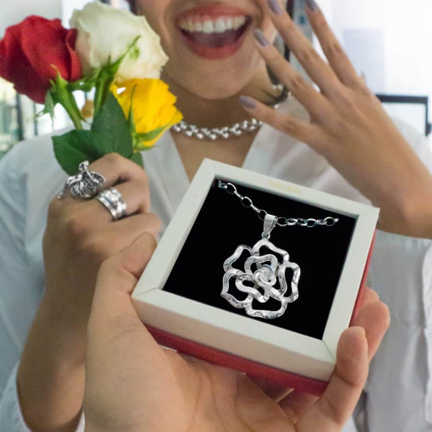 Madre sonriente sosteniendo una caja de regalo con un colgante de plata en forma de flor mientras exhibe orgullosa su anillo y collar de plata, con rosas en la mano