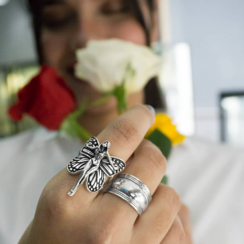 Una mamá enfocando su mano hacia la cámara mostrando un espectacular anillo de plata con diseño de hada, complementado por otros anillos de plata, con un desenfoque artístico de rosas en el fondo
