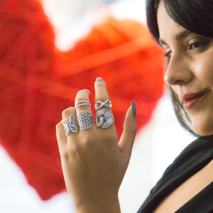 Mano femenina con varios anillos de plata con diseños originales, incluyendo formas de lazo y Flor de la Vida, mariposa, infinito .. ideales para un regalo de San Valentín